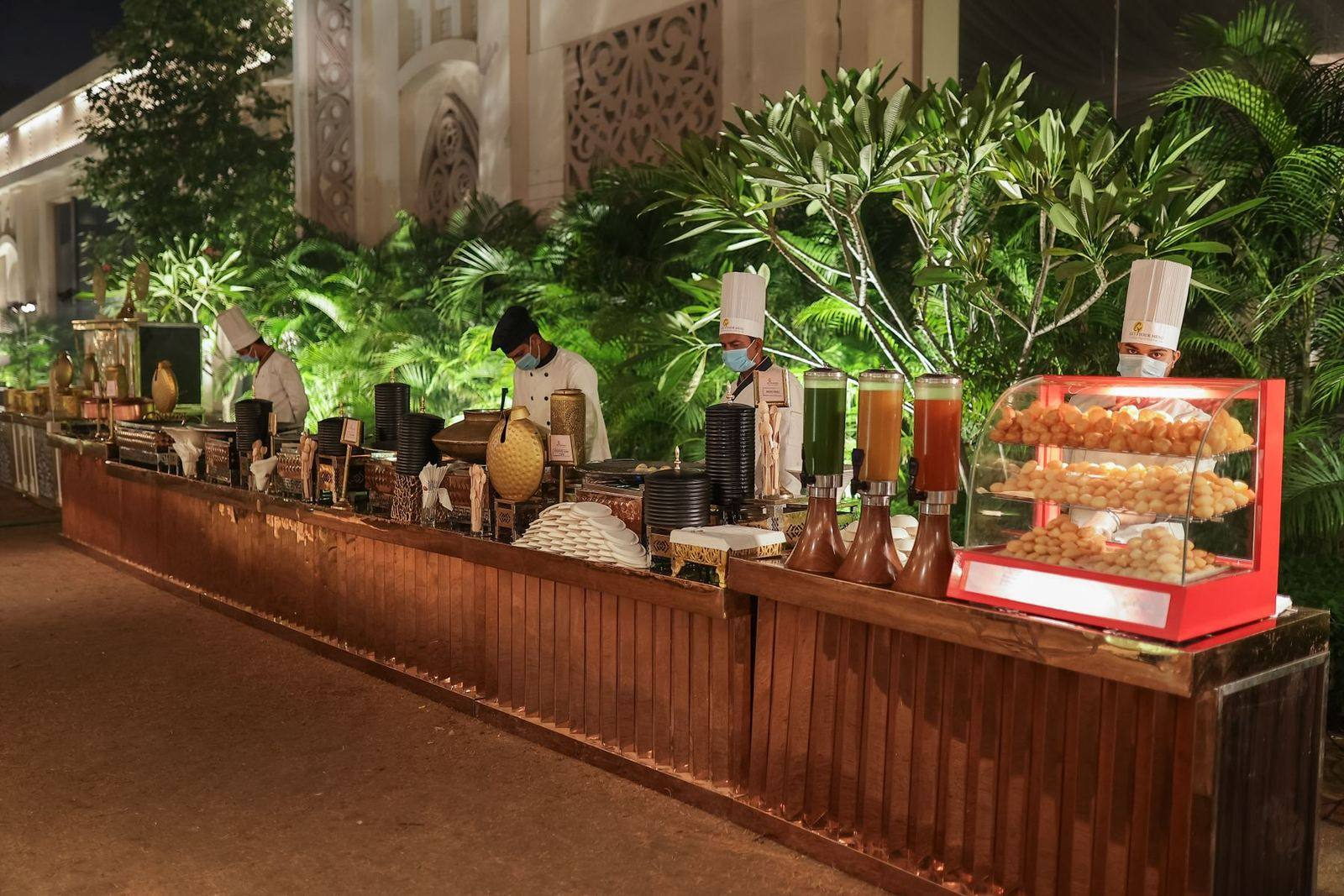 A long outdoor catering counter at night with chefs at their stations, juice dispensers and a lit display case of puris. Illustrative photograph.