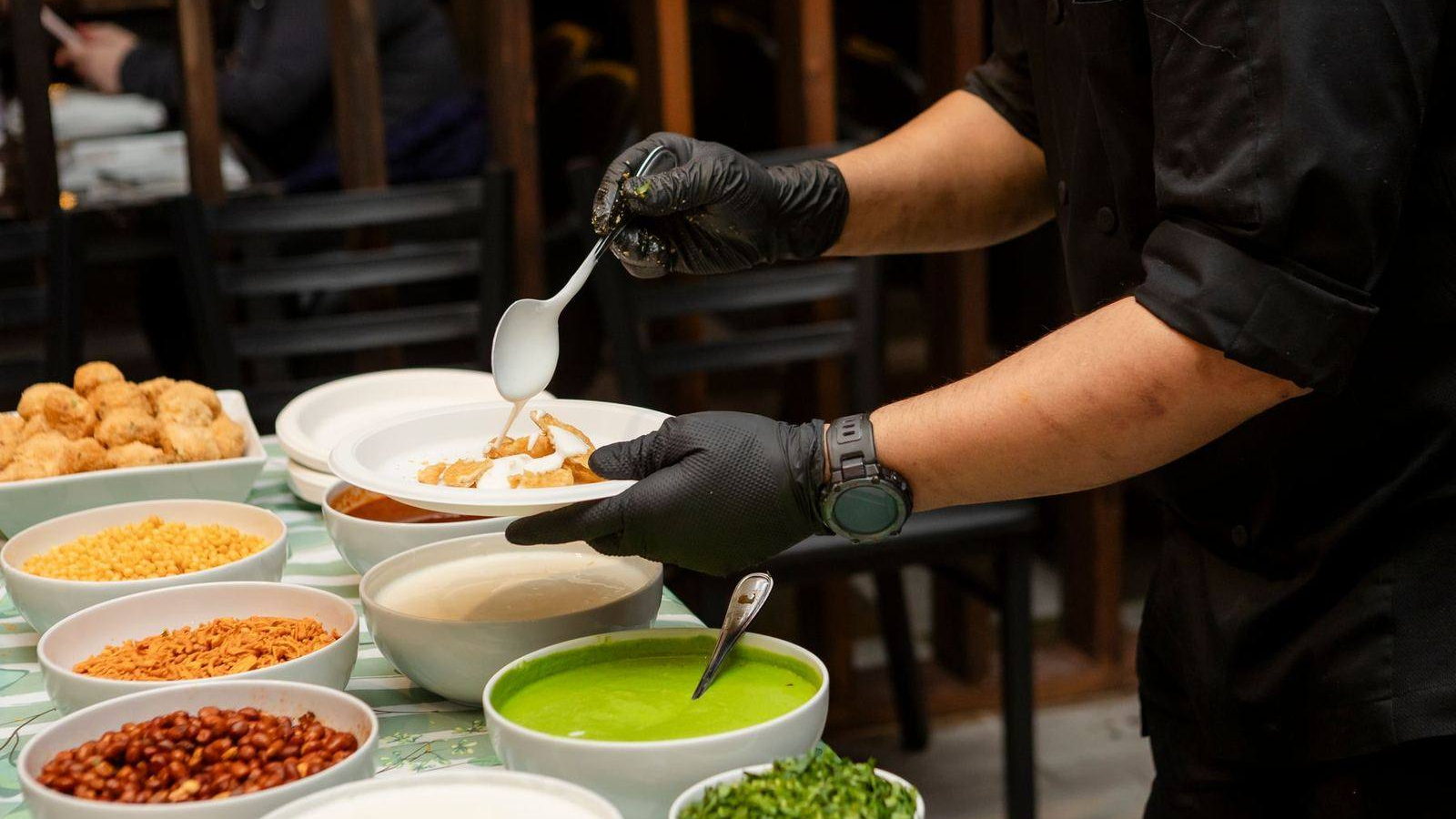 A cook in black gloves spoons curd over a plate of chaat at a counter lined with bowls of sev, peanuts, chutneys and fried puris. Illustrative photograph.