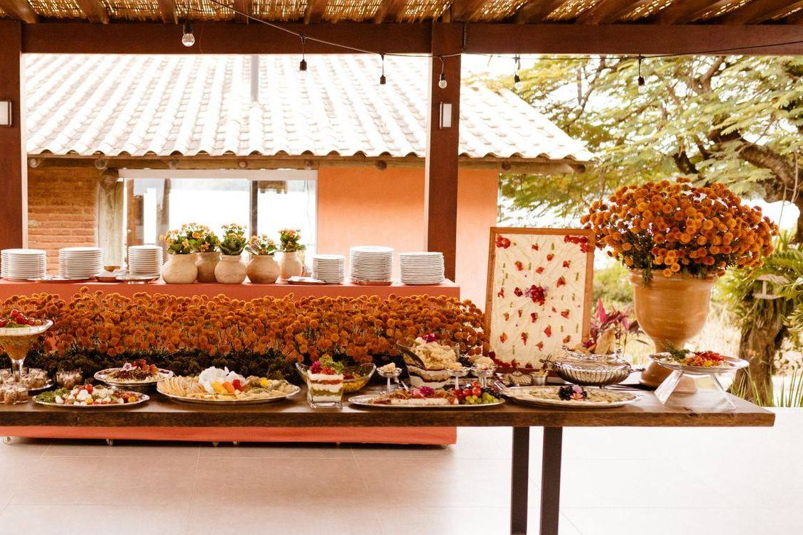 A long buffet table dressed with orange flowers and stacked plates, set with platters of vegetarian dishes under a covered terrace. Illustrative photograph.