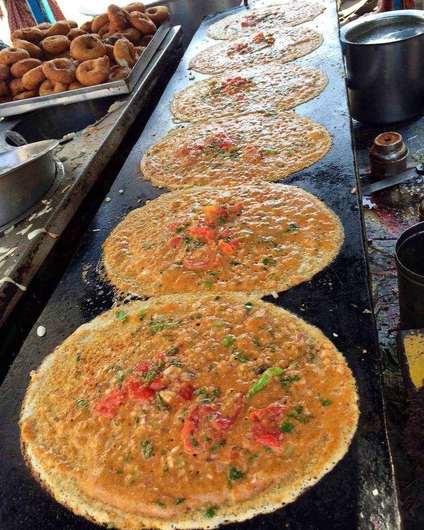 A row of onion-and-tomato uttapam cooking along a long flat griddle, with a tray of fried vadas at the side. Illustrative photograph.