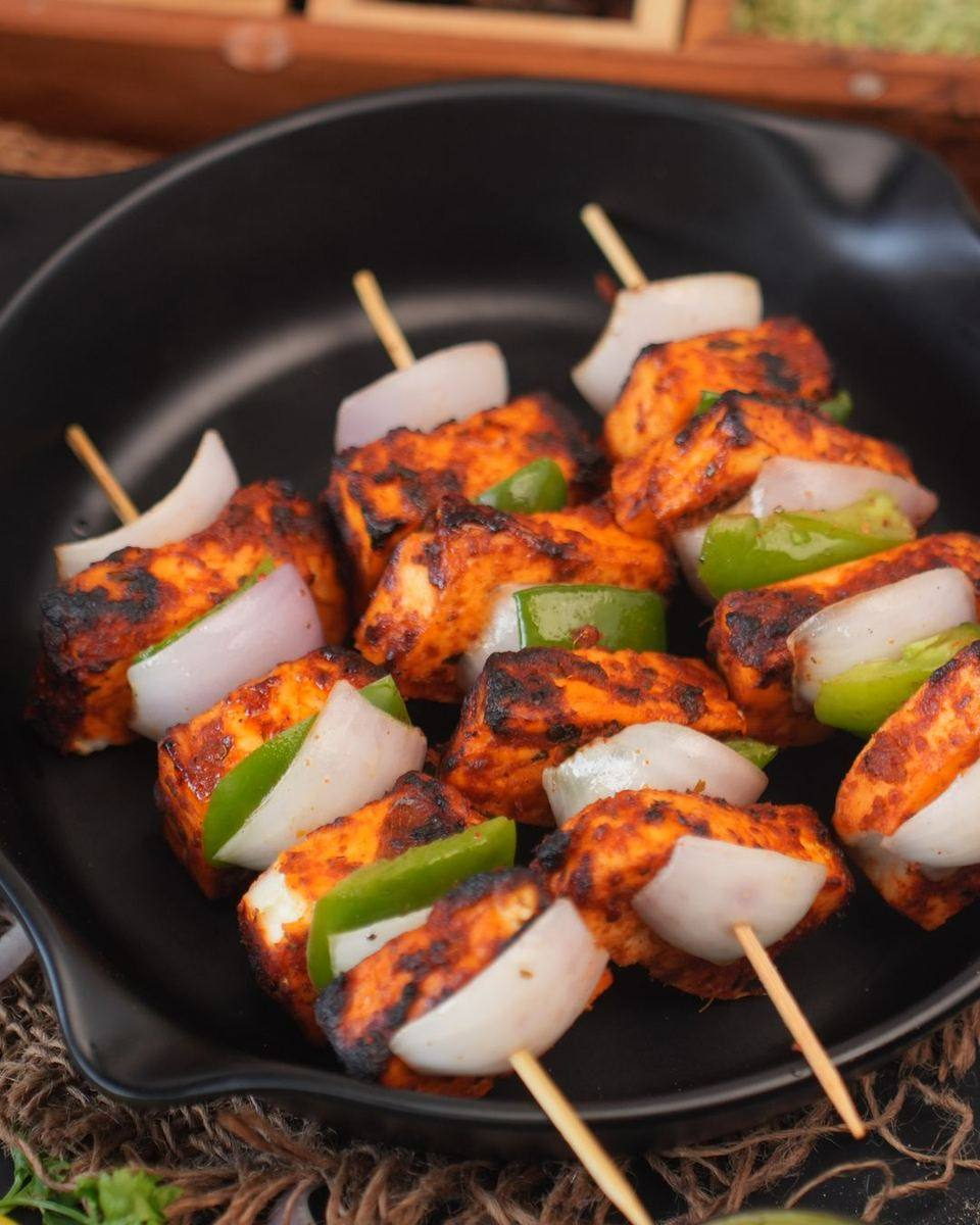Skewers of charred marinated paneer with onion and capsicum resting in a black cast-iron pan. Illustrative photograph.
