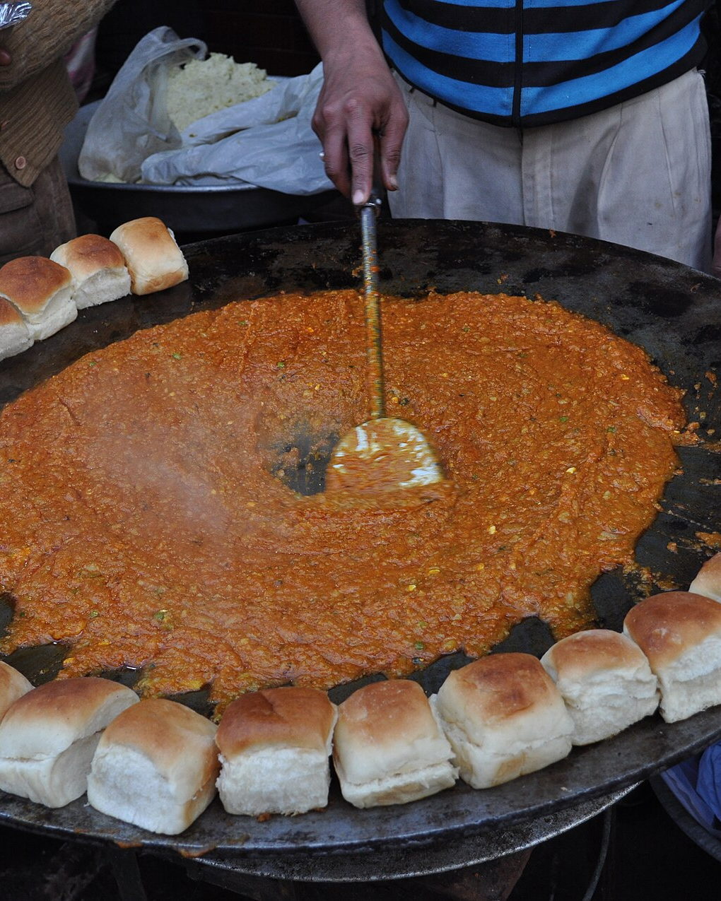 Bhaji simmering across a very large iron tawa ringed with pav buns, a cook's hand working the ladle. Illustrative photograph.