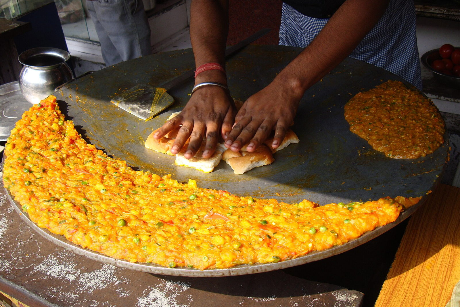 A cook's hands pressing pav onto a large iron tawa beside a ring of cooked bhaji. Illustrative photograph.